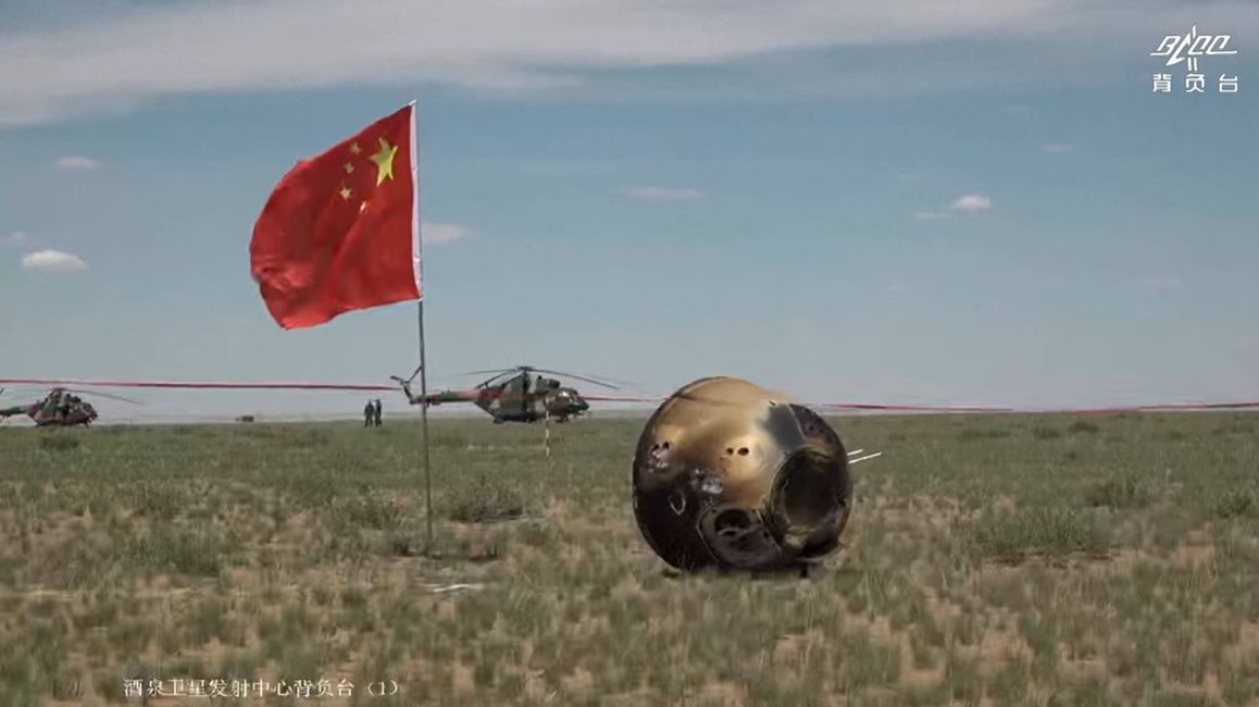 A Chinese flag stands next to a scorched, tipped over Chang'e-6 reentry capsule in the grasslands of Inner Mongolia. Helicopters are visible in the distance.