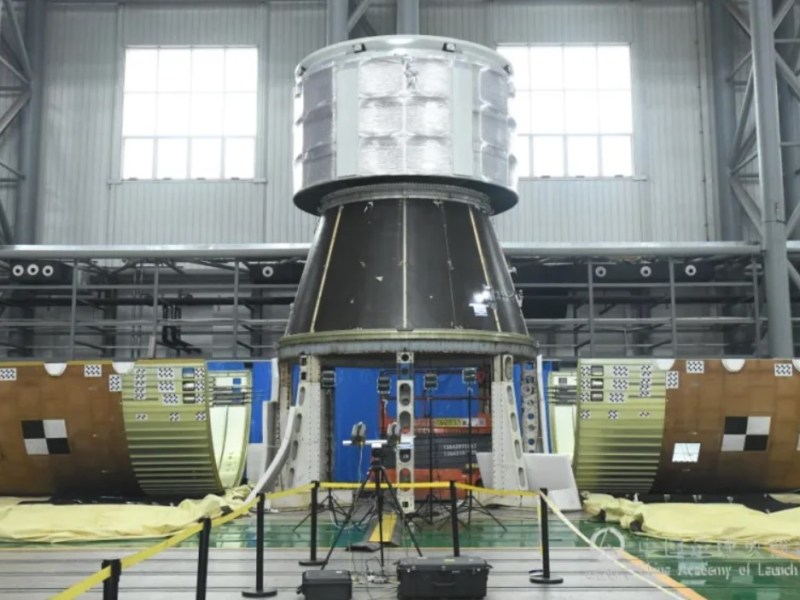 Image showing the payload fairing separation test for the Long March 10 rocket, featuring the separated fairing halves on the ground and the structural test article exposed in the center. The test setup includes a conical spacecraft section with a metallic upper structure, surrounded by a controlled testing environment with scaffolding and observation equipment.
