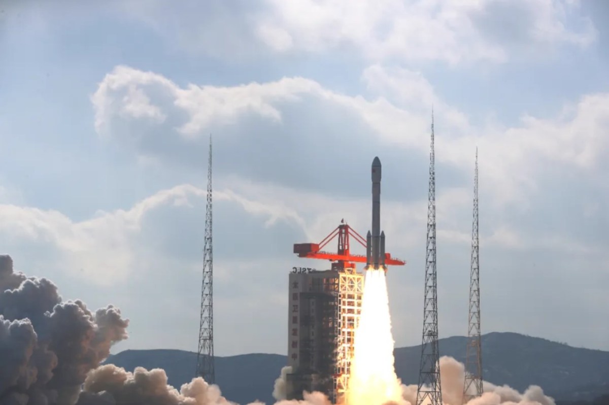 A Long March 6A rocket lifts off from the Taiyuan Satellite Launch Center in northern China, surrounded by four towering lightning rods, leaving a plume of dense white exhaust against a backdrop of a partly cloudy blue sky. The launch marks the deployment of the third batch of 18 Qianfan (Thousand Sails) satellites, a key step in China's ambitious low Earth orbit megaconstellation project, December 5, 2024.
