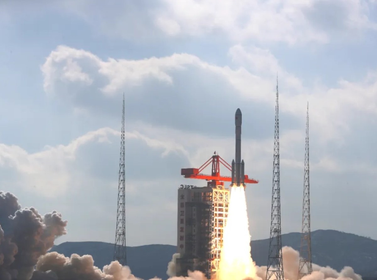 A Long March 6A rocket lifts off from the Taiyuan Satellite Launch Center in northern China, surrounded by four towering lightning rods, leaving a plume of dense white exhaust against a backdrop of a partly cloudy blue sky. The launch marks the deployment of the third batch of 18 Qianfan (Thousand Sails) satellites, a key step in China's ambitious low Earth orbit megaconstellation project, December 5, 2024.