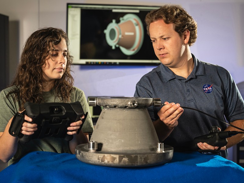 Two NASA engineers examine a 3D-printed aluminum nozzle created under Marshall Space Flight Center’s Reactive Additive Manufacturing for the Fourth Industrial Revolution (RAMFIRE) project. One engineer holds a diagnostic device while the other inspects the nozzle’s surface. In the background, a computer screen displays a 3D model of the component, highlighting the project’s focus on innovation and advanced manufacturing techniques.