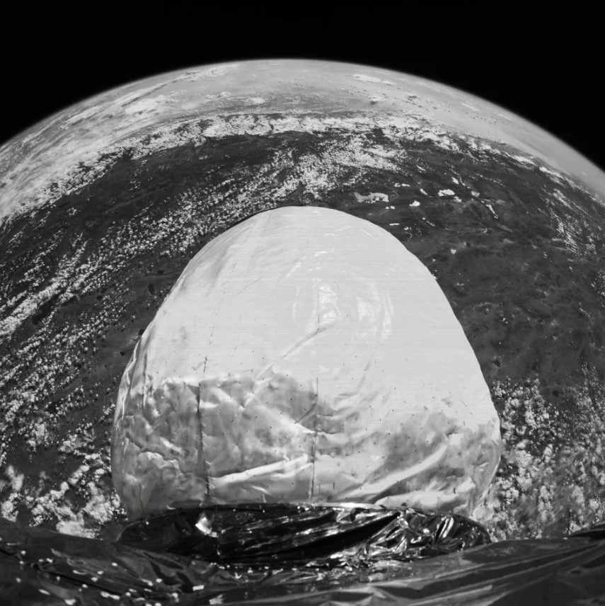 A black-and-white image of an inflatable space habitat module deployed in orbit, with Earth’s surface visible in the background. The module features a smooth, rounded structure and reflective material at its base, showcasing its lightweight and flexible design.