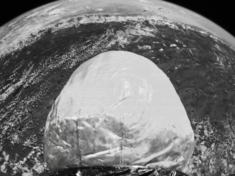 A black-and-white image of an inflatable space habitat module deployed in orbit, with Earth’s surface visible in the background. The module features a smooth, rounded structure and reflective material at its base, showcasing its lightweight and flexible design.