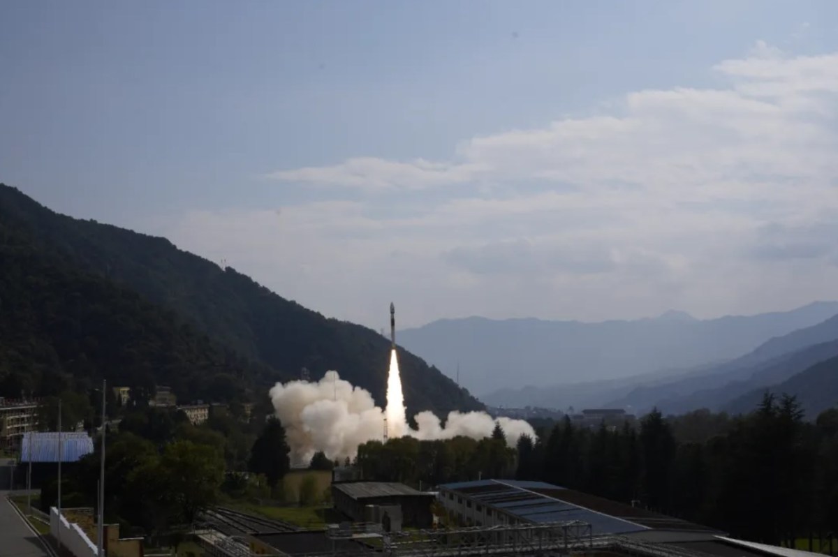 Rocket launch from a mountainous spaceport, showing a solid-propellant rocket ascending against a clear blue sky with smoke trailing from the launch pad, surrounded by forested hills