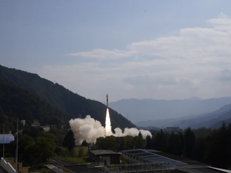 Rocket launch from a mountainous spaceport, showing a solid-propellant rocket ascending against a clear blue sky with smoke trailing from the launch pad, surrounded by forested hills