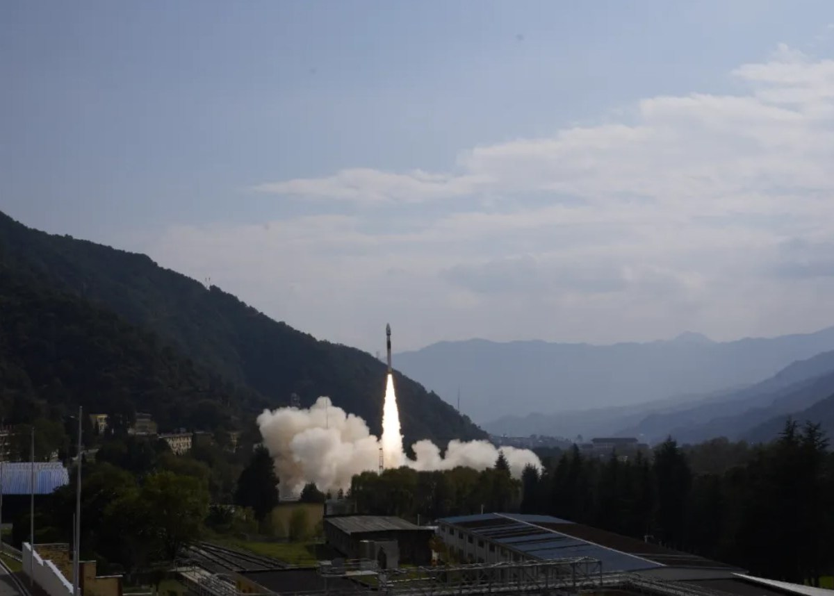 Rocket launch from a mountainous spaceport, showing a solid-propellant rocket ascending against a clear blue sky with smoke trailing from the launch pad, surrounded by forested hills