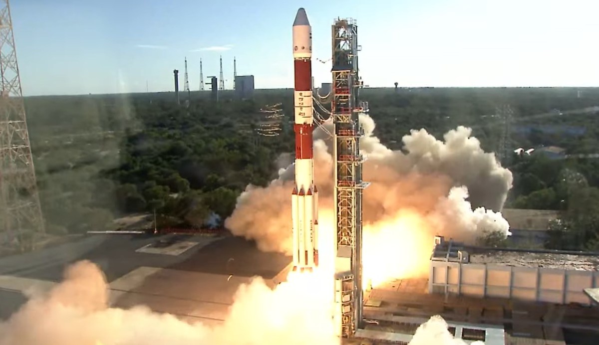 A Polar Satellite Launch Vehicle (PSLV) lifts off from the Satish Dhawan Space Centre, surrounded by smoke and flames, against a backdrop of green vegetation and blue sky. The rocket's red-and-white design stands out as it carries the European Proba-3 mission into orbit.