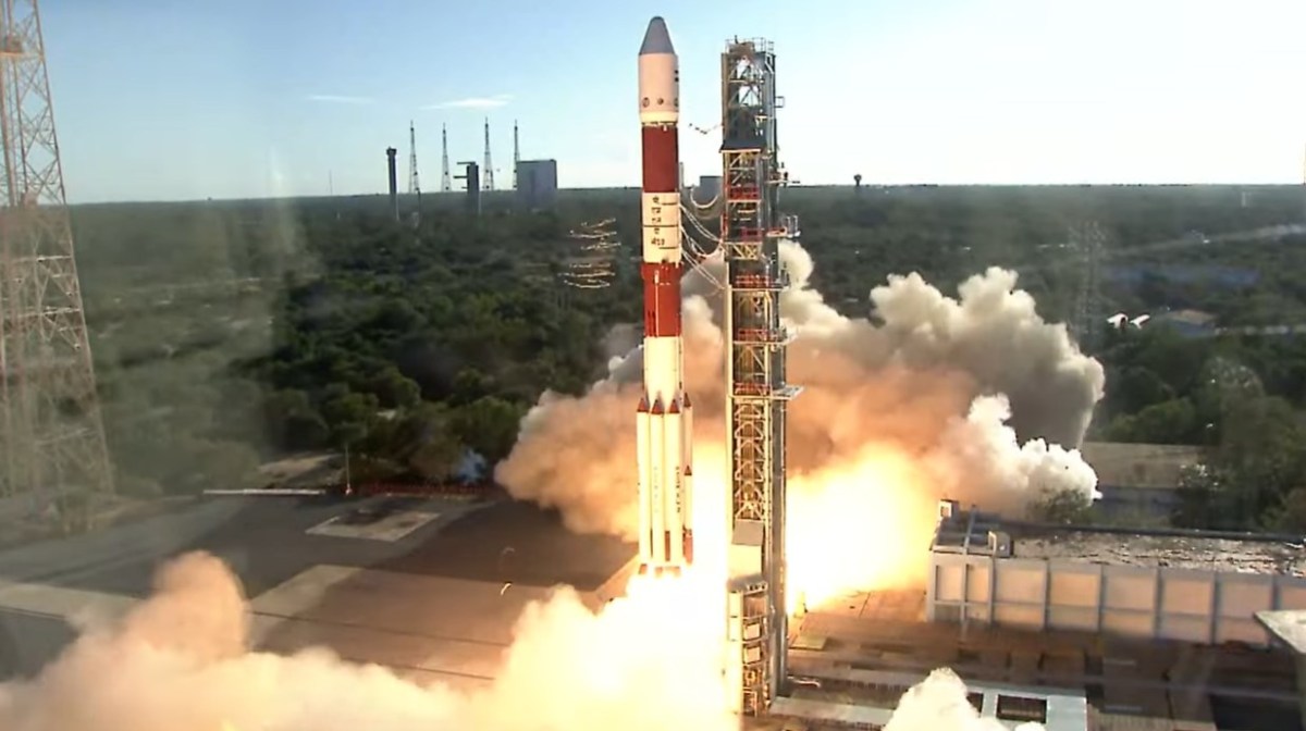 A Polar Satellite Launch Vehicle (PSLV) lifts off from the Satish Dhawan Space Centre, surrounded by smoke and flames, against a backdrop of green vegetation and blue sky. The rocket's red-and-white design stands out as it carries the European Proba-3 mission into orbit.