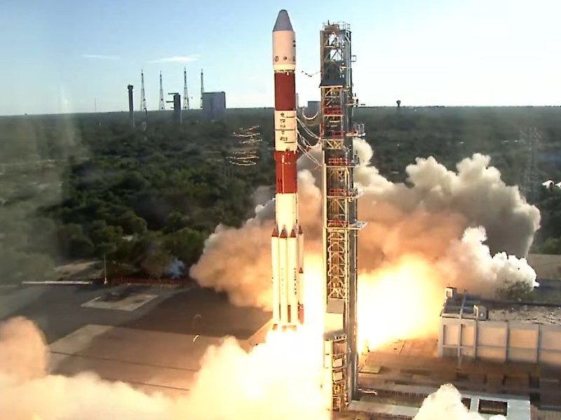 A Polar Satellite Launch Vehicle (PSLV) lifts off from the Satish Dhawan Space Centre, surrounded by smoke and flames, against a backdrop of green vegetation and blue sky. The rocket's red-and-white design stands out as it carries the European Proba-3 mission into orbit.