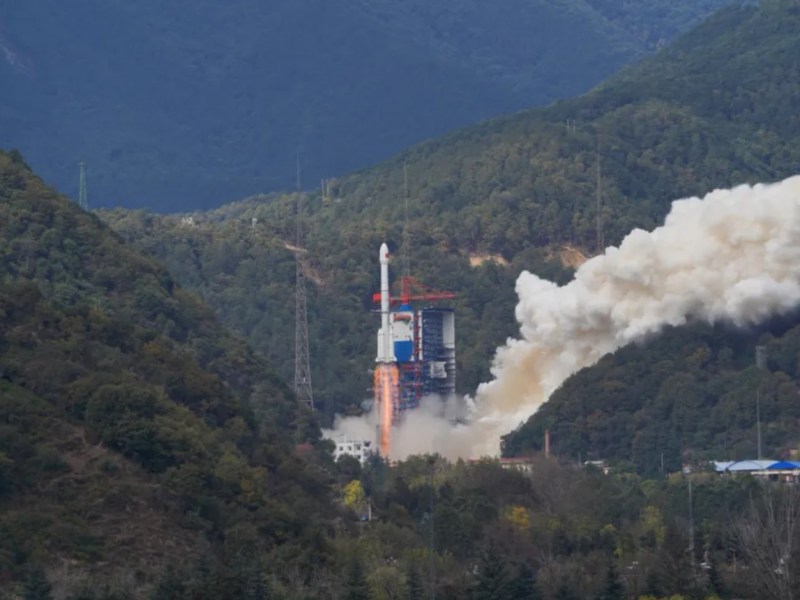 A Long March 3B rocket lifts off from the Xichang Satellite Launch Center in a verdant, mountainous region, leaving a trail of smoke, marking its 100th flight and carrying the TJS-13 satellite into orbit.
