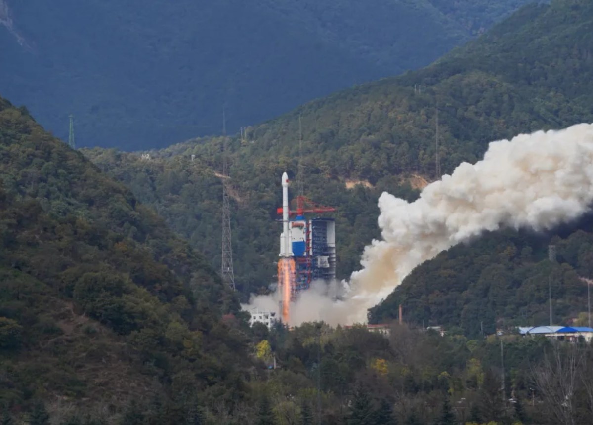 A Long March 3B rocket lifts off from the Xichang Satellite Launch Center in a verdant, mountainous region, leaving a trail of smoke, marking its 100th flight and carrying the TJS-13 satellite into orbit.