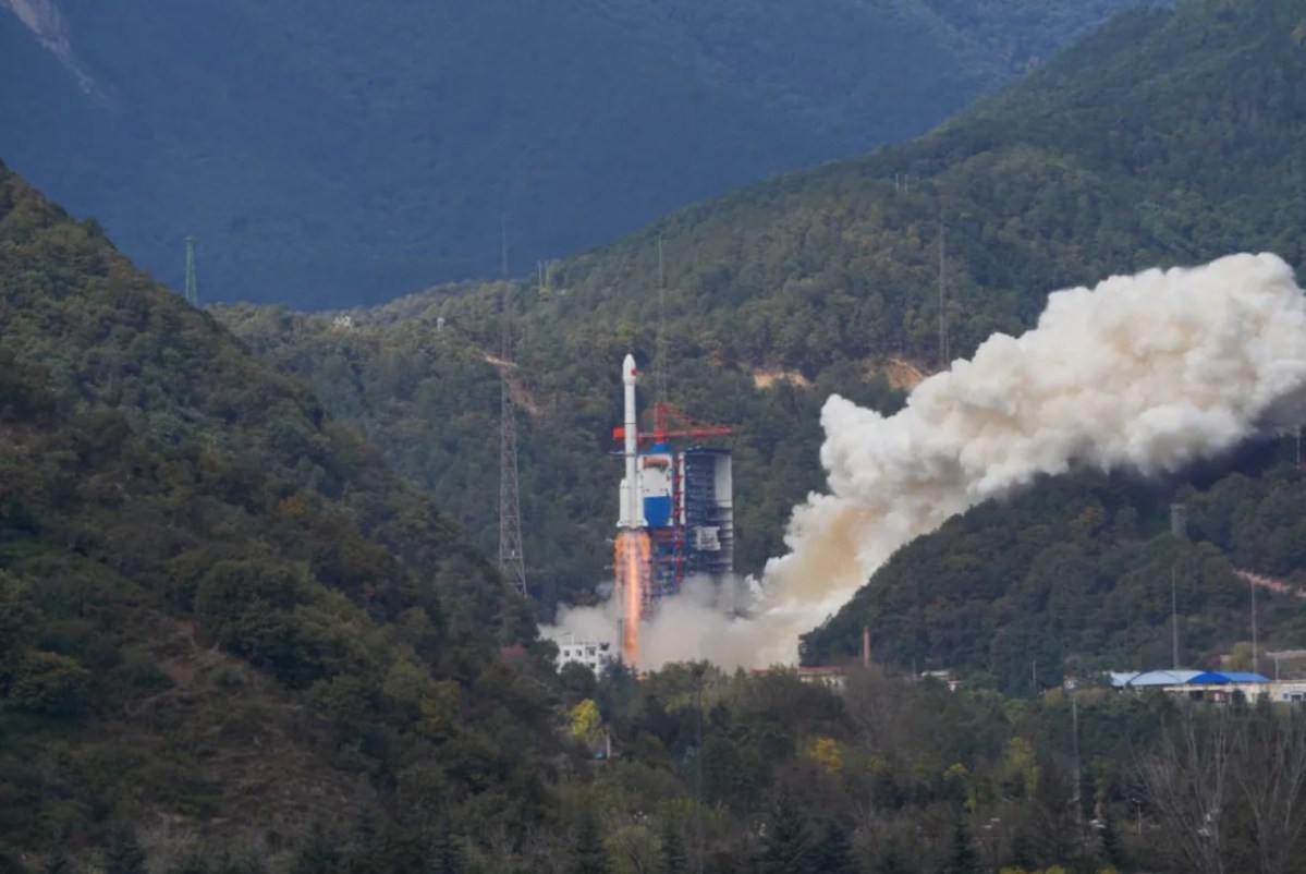 A Long March 3B rocket lifts off from the Xichang Satellite Launch Center in a verdant, mountainous region, leaving a trail of smoke, marking its 100th flight and carrying the TJS-13 satellite into orbit.