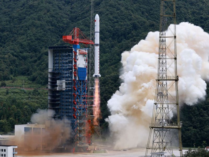 A 41-meter-long white Long March 2D rocket rises from its launch tower at hill-surrounded Xichang launch center. Mach diamonds form in the exhaust while plumes of white smoke billow away from the path of the rocket.
